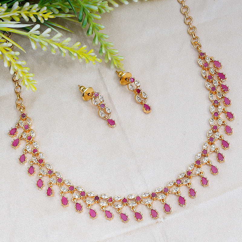 A gold necklace and matching earrings with pink and white gemstones, displayed on a light fabric background with green foliage.