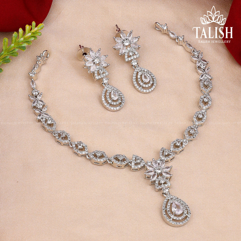 A silver necklace and earring set with intricate designs and sparkling gemstones, displayed on a beige background.