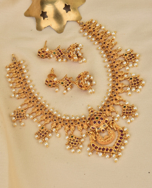 A set of traditional gold jewelry featuring a necklace and matching earrings adorned with intricate designs, red gemstones, and small white pearls.