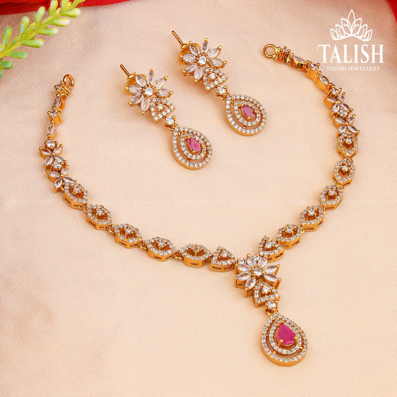 A gold necklace and earring set with intricate designs, featuring clear and pink gemstones, displayed on a soft fabric background.