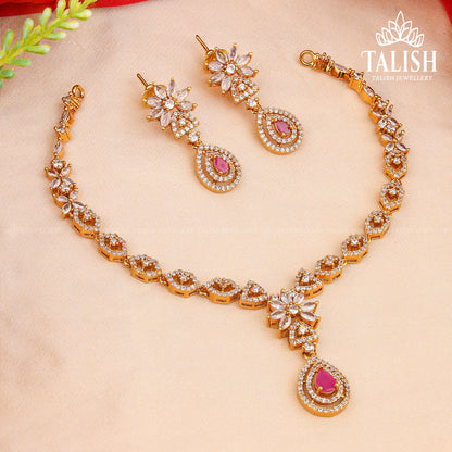 A gold necklace and earring set with intricate designs, featuring clear and pink gemstones, displayed on a soft fabric background.