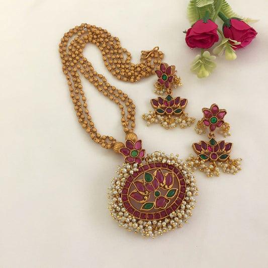 A traditional gold necklace with a large, round pendant featuring red and green floral designs, accompanied by matching earrings.