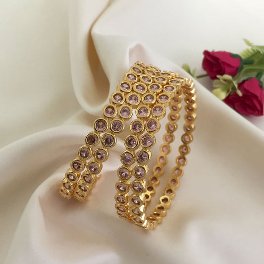 Two gold bangles adorned with pink gemstones placed on a white fabric.