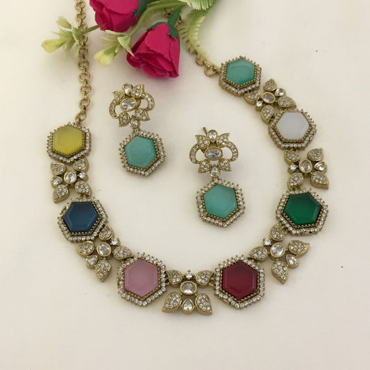 A gold necklace and earring set with hexagonal multicolored gemstones and diamond accents. The necklace features a chain and a series of hexagonal stones in various colors, including yellow, blue, pink, green, and white, surrounded by small diamonds. The matching earrings have a similar design with a central hexagonal stone and diamond accents.