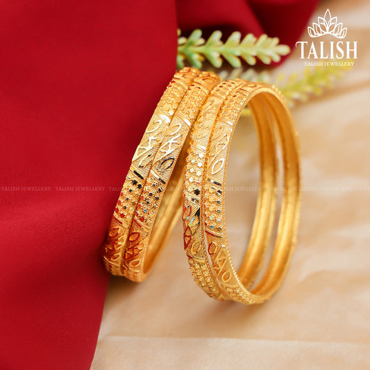 A pair of intricately designed gold bangles on a red fabric background with a small green plant.