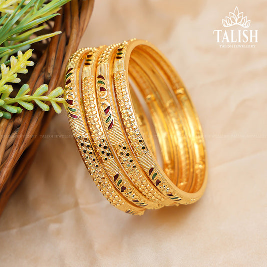 A set of intricately designed gold bangles with colorful enamel details, placed on a beige surface next to a wicker basket with green foliage.