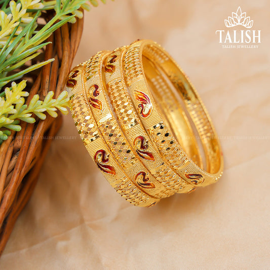 A set of intricately designed gold bangles with red and black enamel accents, placed on a beige surface next to a wicker basket and green foliage.