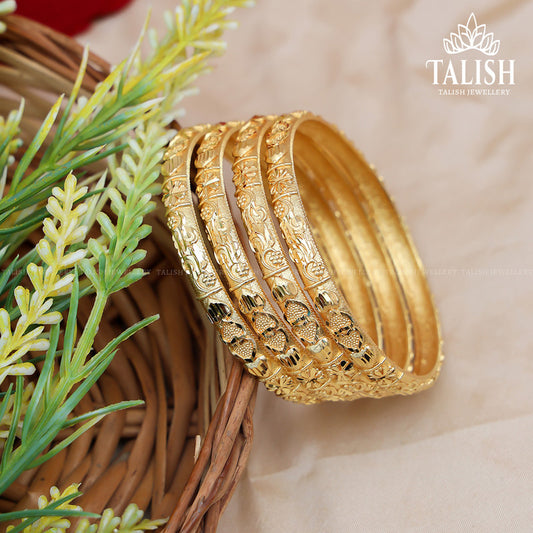 A set of intricately designed gold bangles placed in a woven basket with green foliage nearby.