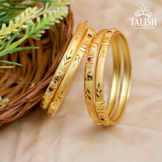 A set of intricately designed gold bangles with red gemstone accents, placed next to a woven basket and green foliage.