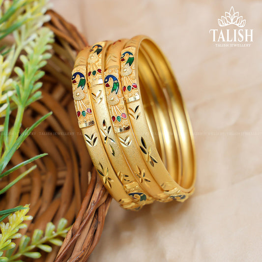 A set of intricately designed gold bangles with colorful enamel detailing, placed in a wicker basket next to green foliage.