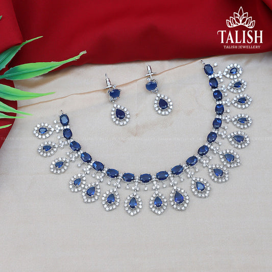 A set of elegant jewelry featuring a necklace and matching earrings with blue gemstones and silver accents, displayed on a beige surface with a red fabric background.