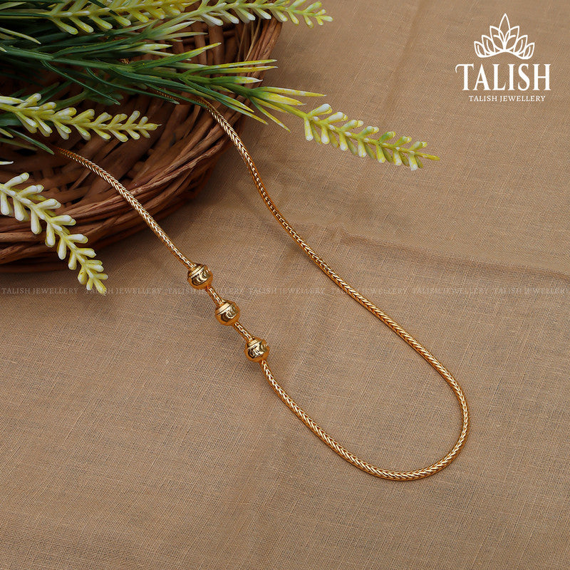 A gold necklace with three decorative beads displayed on a brown fabric surface, next to a basket with green foliage.