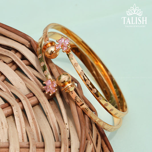 Two gold bangles with intricate designs and pink floral embellishments, placed on a woven basket surface.