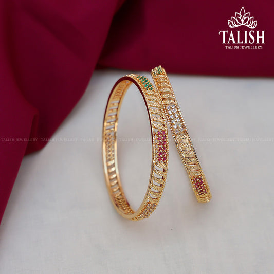 A pair of intricately designed gold bangles with red, green, and white gemstones, displayed on a white surface with a maroon fabric background.