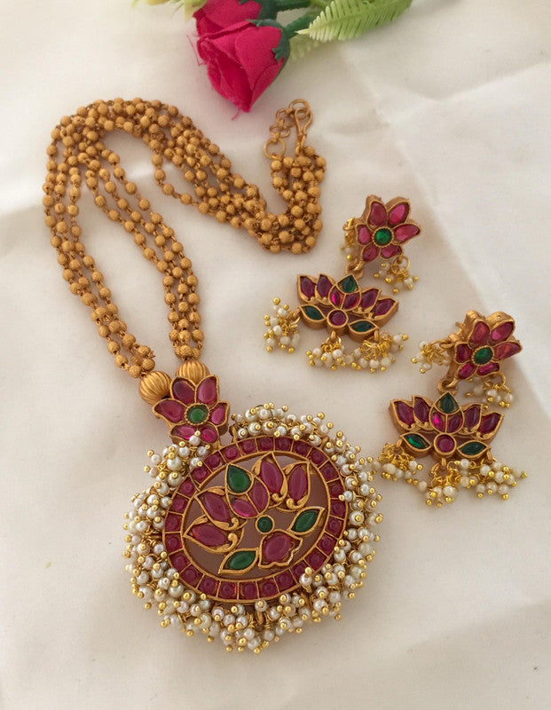 A traditional Indian jewelry set featuring a necklace with a large, ornate pendant and matching earrings. The design includes red and green stones with intricate gold detailing and pearl accents.