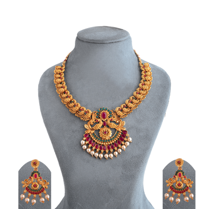 Gold necklace with intricate design, featuring red and green gemstones and pearl accents, displayed on a gray stand.