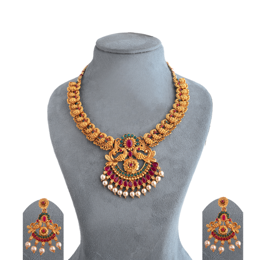 Gold necklace with intricate design, featuring red and green gemstones and pearl accents, displayed on a gray stand.