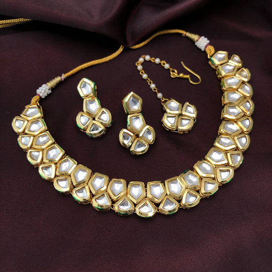 A gold necklace set with white gemstones, including a necklace, a pair of earrings, and a maang tikka, displayed on a dark fabric background.