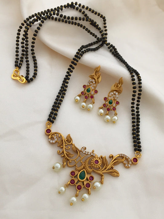 A traditional gold-toned mangalsutra set with black beads, featuring an intricate pendant adorned with red, green, and white stones, and matching earrings.