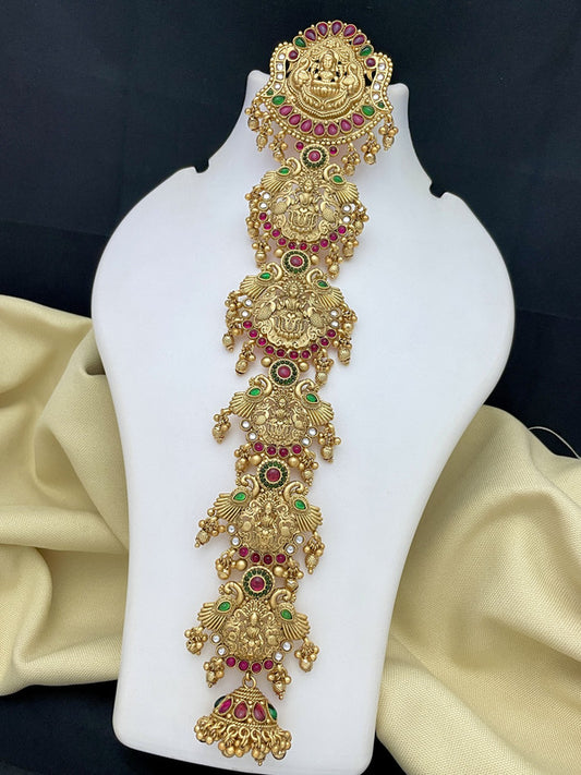 Gold temple jewelry with intricate designs, adorned with red and green gemstones, displayed on a white stand.