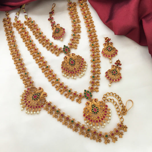 A set of traditional gold jewelry with intricate designs, featuring necklaces, earrings, and a maang tikka adorned with red and green gemstones and pearl accents.