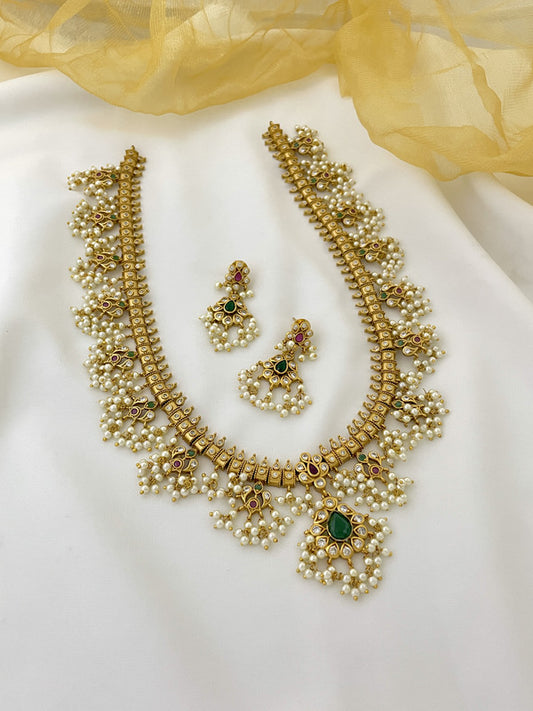 A gold necklace with intricate design and pearl embellishments, accompanied by matching earrings, displayed on a white surface.