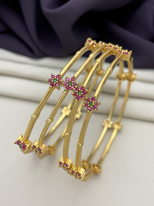 Gold bangles with floral designs adorned with pink and green gemstones.