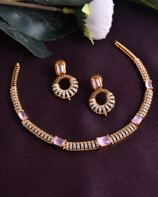 A gold necklace and matching earrings set with pink and white stones displayed on a dark surface.