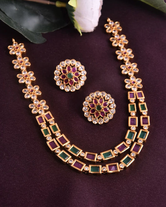 A gold necklace with intricate floral designs and matching earrings, featuring red and green gemstones.