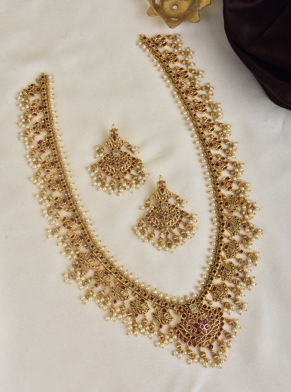 A traditional gold necklace set with intricate designs and matching earrings, adorned with small pearls and red gemstones, displayed on a white surface.