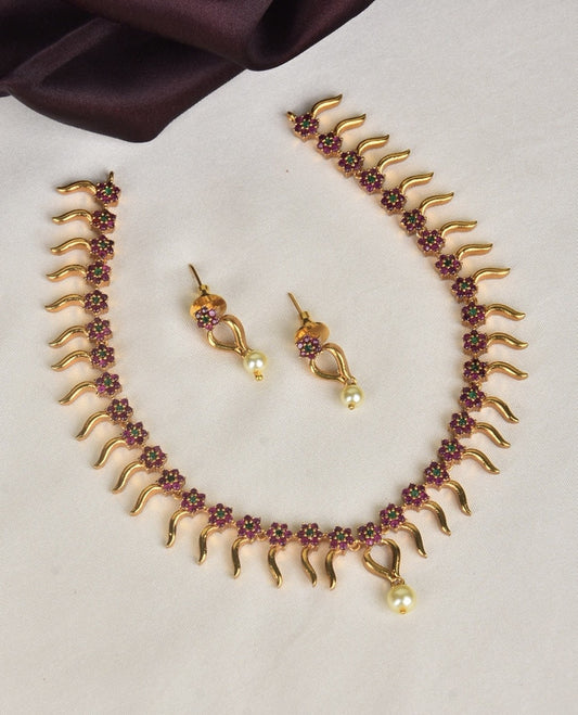 A gold necklace with a wavy design, adorned with small red flowers and matching earrings, each featuring a red flower and a dangling pearl.