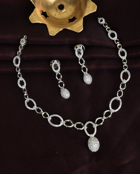 A silver jewelry set consisting of a necklace and matching earrings, featuring oval-shaped links and teardrop-shaped pendants encrusted with small crystals.