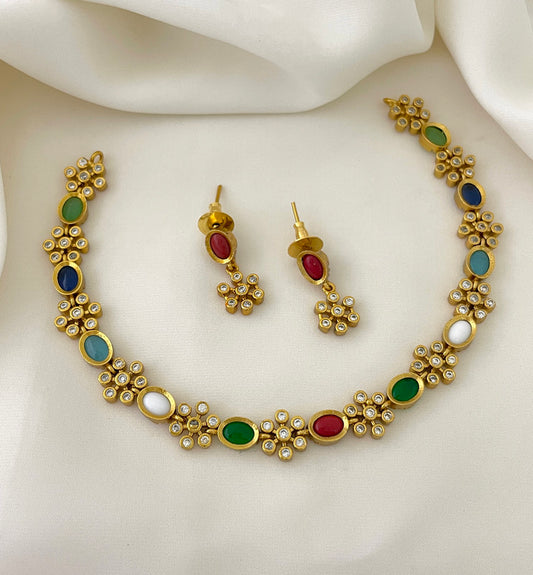 A gold jewelry set featuring a necklace and a pair of earrings. The necklace has a floral design with multicolored stones, including blue, green, red, and white. The earrings match the necklace with red stones and floral accents.