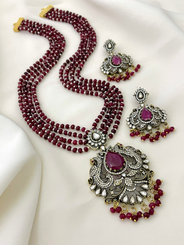 A set of traditional jewelry featuring a multi-strand red beaded necklace with an ornate pendant adorned with a large central red stone, smaller white stones, and intricate metalwork. Matching earrings with similar design elements are placed beside the necklace.