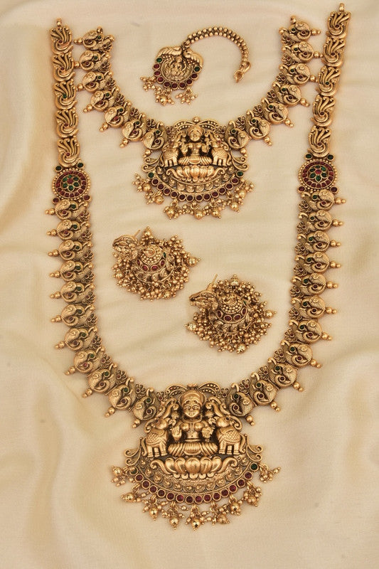 A set of traditional gold jewelry featuring intricate designs, including a necklace, a pair of earrings, and a maang tikka, all adorned with red and green gemstones.