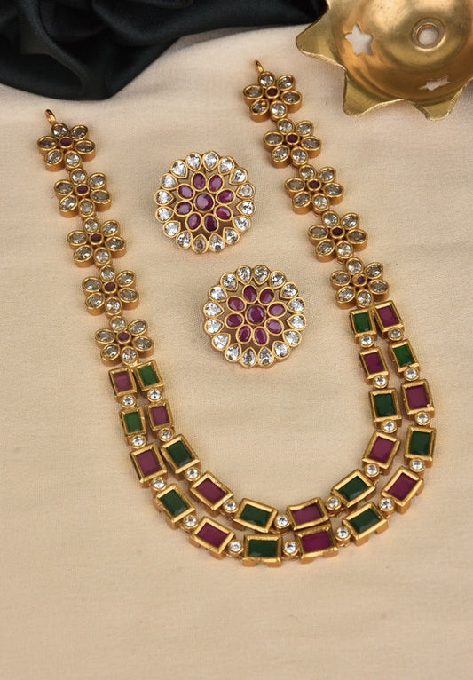 A gold necklace with floral designs and square gemstones in green and purple, accompanied by a pair of matching round earrings with purple and white stones.