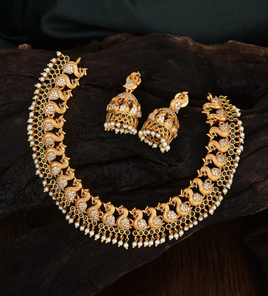 A gold necklace with intricate peacock designs and matching gold earrings. The necklace features detailed craftsmanship with embedded stones and pearl accents. The earrings are designed in a similar style with peacock motifs and dangling pearls.