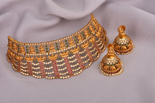 A gold choker necklace with intricate designs and matching jhumka earrings, adorned with green and red gemstones.