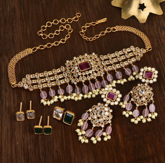 A set of gold jewelry featuring a choker necklace, a pair of chandelier earrings, and two pairs of stud earrings. The choker and chandelier earrings are adorned with clear and pink gemstones, with a central red gemstone. The stud earrings include one pair with green gemstones and another with clear gemstones.