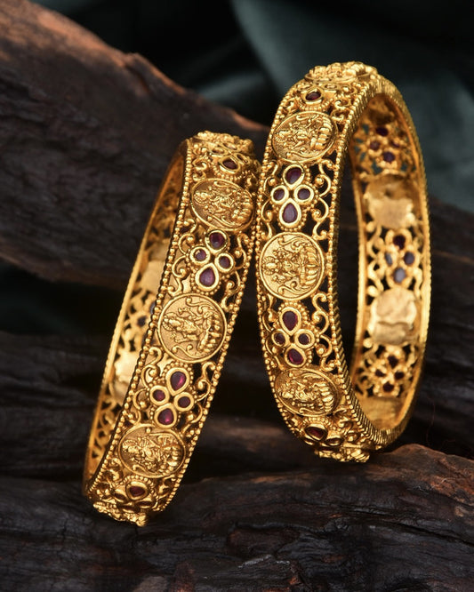 Two intricately designed gold bangles with detailed engravings and embedded red gemstones.