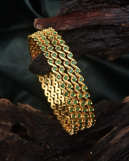 A gold bracelet with an intricate, wavy design and embedded green gemstones.