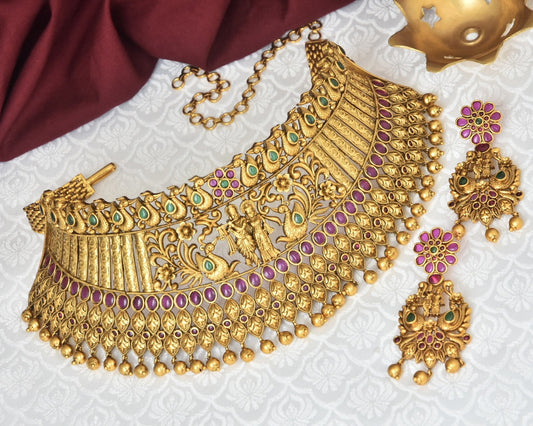 A set of ornate gold jewelry featuring a choker necklace with intricate designs, green and purple gemstones, and matching dangling earrings.