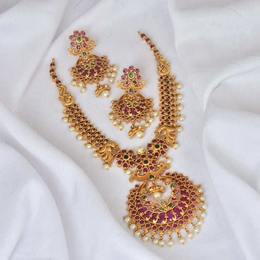 A gold necklace set with intricate designs, featuring red and green gemstones, and matching earrings displayed on a white fabric.