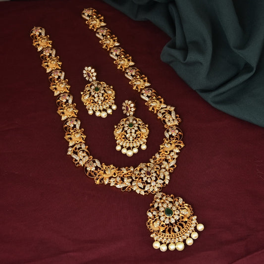 A set of ornate gold jewelry consisting of a long necklace, a pair of earrings, and a maang tikka, all adorned with intricate designs and embedded with gemstones and pearls.