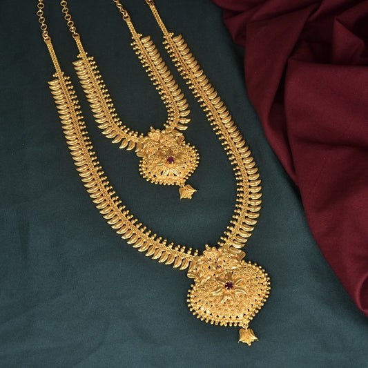 Two intricately designed gold necklaces with ornate pendants, displayed on a dark green surface with a maroon fabric in the background.